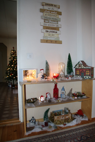 Olohuoneen yhdellä seinustalla on monenlaisia koristeita siistissä järjestyksessä. Yläpuolella on kodin ihanuutta ylistäviä tunnuslauseita. Eteisessä näkyy yksi joulukuusista.